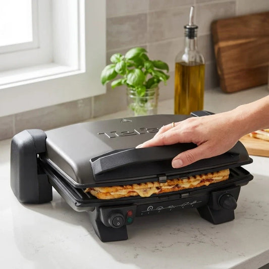 Person using a Tefal grill on a kitchen counter with a plant and bottle in the background.